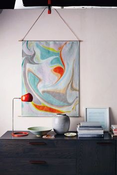 an abstract painting hangs on the wall above a dresser with plates, bowls and a lamp