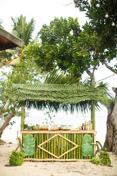 an outdoor bar made out of bamboo sticks and palm leaves on the beach with water in the background