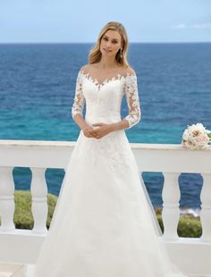 a woman in a white wedding dress standing on a balcony near the ocean with her hand on her hip