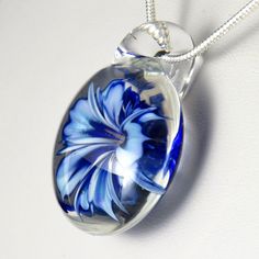 This unusual and unique pendant has a realistic blue and white glass 3D flower inside a solid glass - this isn't resin or a real flower, it's all made entirely of glass! I've used a cobalt blue over white, which I've graduated in tone and to form bands on the petals to make the flower look more realistic, also giving the edges and centre a darker rim of blue. In the centre I've added some yellow 'stamens' to make it look more realistic, where some bubbles have clustered around them to add a dew White Flower Shaped Glass Jewelry, Blue Glass Flower Shaped Jewelry, Blue Flower Shaped Glass Jewelry, Blue Flower-shaped Glass Jewelry, Silver Glass Flower Pendant Jewelry, Blue Round Pendant Jewelry With Flower Charm, Blue Flower Charm Round Pendant Jewelry, Blue Flower Pendant Necklace For Gift, Blue Flower Pendant Necklace With Large Pendant