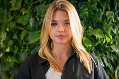 a blonde woman standing in front of a green wall with leaves on it's sides