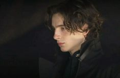 a young man with curly hair wearing a black coat and dark clothes looking off into the distance