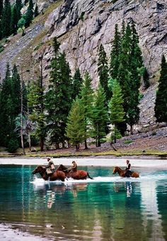 three people are riding horses in the water near some rocks and trees, while another person is on horseback