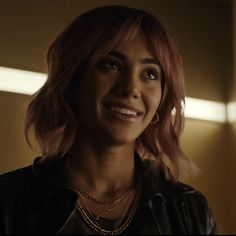 a woman with pink hair wearing a black leather jacket and gold necklace in a dimly lit room