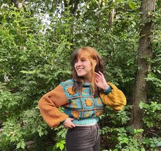 a woman standing in front of trees wearing a colorful sweater and smiling at the camera