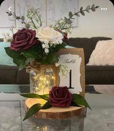 Burgundy and white flower centerpiece. The flowers are placed partially in a jar that is filled with glowing string lights. The entire jar sits on a wooden circle Slab. Mason Jar With Lights Centerpiece, Wedding Table Centerpieces Fairy Lights, Simple Wedding Table Decor Reception Ideas Mason Jars, Simple Rustic Wedding Decorations Table, Mason Jar And Wood Centerpieces, Mason Jar With Fairy Lights And Flowers, Wood Plank Centerpiece, Burlap Centerpieces Table Decorations, Rustic Wedding Centerpieces Mason Jars Fairy Lights