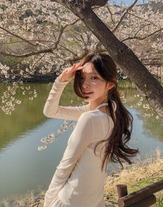 a woman standing next to a tree in front of a lake with cherry blossoms on it