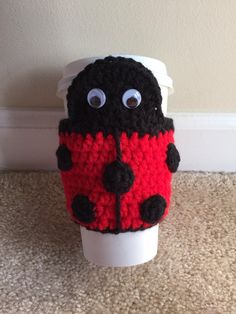 a crocheted coffee cup cozyie with eyes and nose sitting on the floor