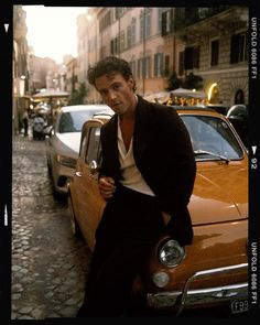 a man leaning on the hood of a car in an old european street with parked cars