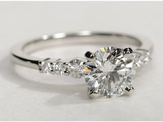 a white gold engagement ring with three stones on the band and a round brilliant cut diamond in the center