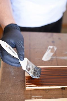 a person in black gloves holding a paintbrush over a piece of wood with brown stain on it