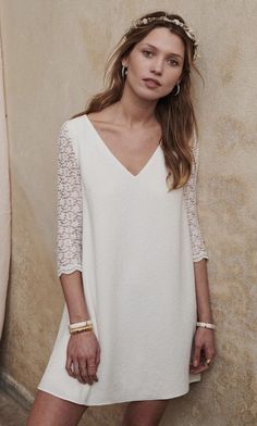 a woman standing in front of a wall wearing a white dress and gold bracelets