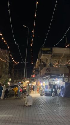 people are sitting on the sidewalk in front of some shops at night with lights strung over them