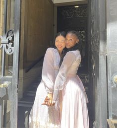 two women in long dresses are standing on the steps of a building and hugging each other