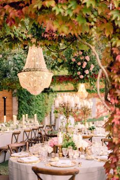 an outdoor dinner table with chandelier hanging from the ceiling and greenery around it