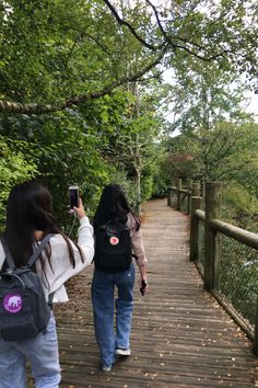 two women walking down a wooden path holding cell phones in their hands and taking pictures