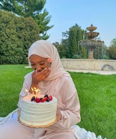 a woman sitting in the grass with a cake on her lap and blowing out candles