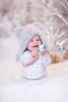 a baby is wearing a furry hat and holding a christmas ornament in the snow