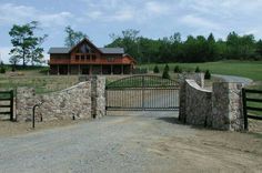 a stone fence and gate in front of a large house on a dirt road with trees around it