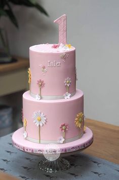 a three tiered pink cake with daisies on the bottom and one number 1 on top