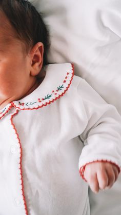 a baby laying on top of a bed wearing a white shirt and red trimming