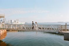 two people walking across a bridge over water near a boat dock with boats in the background