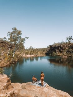 two naked people sitting on rocks near a lake