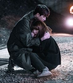 a man and woman sitting on the ground in front of a car with their arms around each other