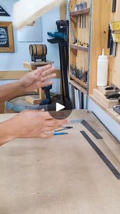 two hands are working on a piece of wood with tools in the background, while another person holds out their hand