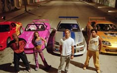 three men and two women standing in front of four different colored cars on the street