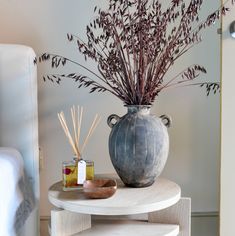 a white table topped with a vase filled with purple flowers and reeds next to a bed