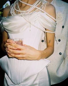 a woman in a white dress and pearls holding a glass with her arm wrapped around her chest