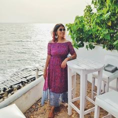 a woman standing next to a white table near the ocean with chairs and a tree in front of her