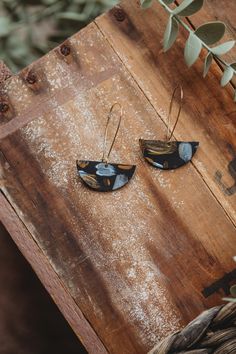 two small black and gold earrings on top of a wooden table next to a plant