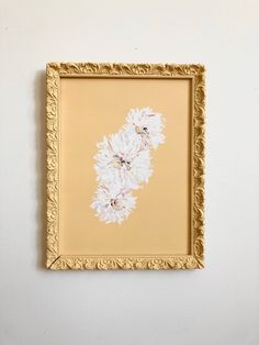 a painting hanging on the wall with white flowers painted on it's gold frame