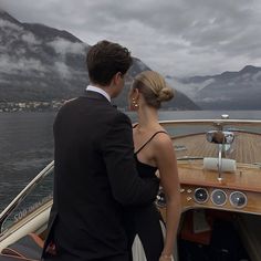 a man and woman standing on the back of a boat looking out over water with mountains in the background