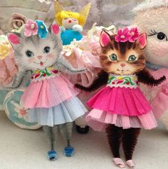 three stuffed animals are dressed in pink and blue tutu skirts, one cat is wearing a flowered dress