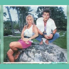 a man and woman sitting on top of a rock next to each other in front of palm trees