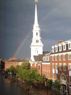 a church steeple with a rainbow in the sky above it and buildings on both sides