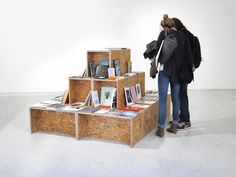 a woman standing next to a stack of books on top of a wooden shelf in front of a white wall