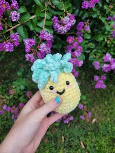 a hand holding a small crocheted toy in front of purple flowers