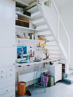 there is a desk under the stairs in this small room with white walls and flooring