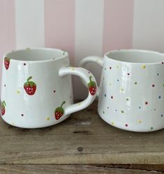 two white mugs with strawberries on them sitting on a wooden table next to a striped wall