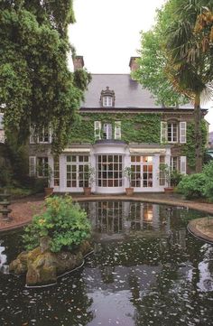 a pond in front of a large house with lots of trees and plants around it