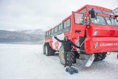 a man standing in front of a red bus on snow covered ground with mountains in the background