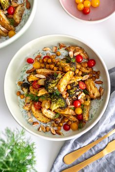 two bowls filled with vegetables and chickpeas on top of a white table next to utensils