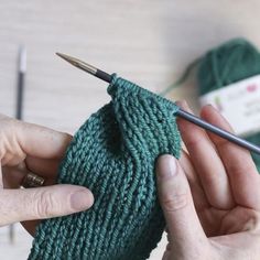 two hands holding yarn and knitting needles in front of a green knitted sock with the needle pointed at the end