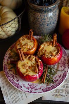 three apples with cinnamon stick toppings on a purple and white plate next to other fruit