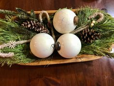 three white balls and pine cones are in a wooden bowl on a table with greenery