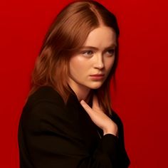 a woman with red hair is posing in front of a red background and she has her hand on her chin